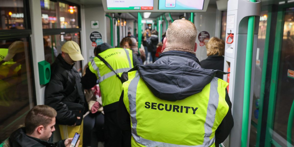 Zwei Mitarbeiter des Sicherheitsdienstes laufen durch eine volle Straßenbahn. Mit ihren hellgelben Westen, auf dneen "Security" steht, sind sie gut zu erkennen.